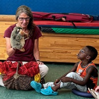Petra Kern sitzt mit einer Igel-Handpuppe zwischen zwei Kindern, die sie interessiert anschauen.