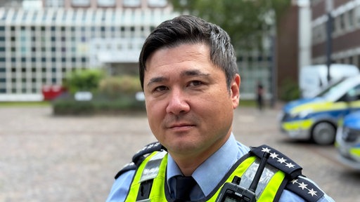 Ein Polizist steht vor der Polizeipräsidium in Dortmund und schaut in die Kamera.