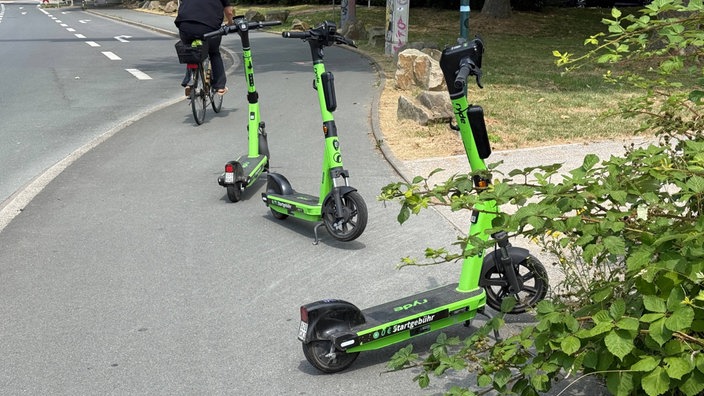 Drei geparkte E-Scooter stehen auf einem Gehweg, links fährt eine Radfahrerin an ihnen vorbei.