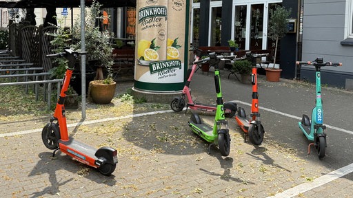 Mehrere E-Scooter stehen auf einem dafür gekennzeichneten Parkplatz, eine Litfaßsäule ist im Hintergrund.