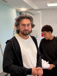 Ein Mann mit schwarzer Kapuzenjacke und weißem T-Shirt blickt in die Kamera, hinter ihm sind Schülerinnen und Schüler in einem Klassenzimmer
