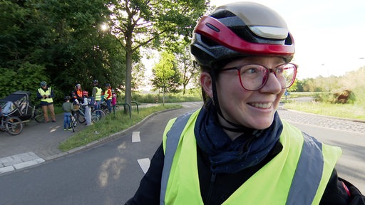 Eine Frau mit rotem Fahrradhelm und gelber Warnweste steht an einem Kreisel, im Hintergrund sind Kinder und Eltern mit ihren Fahrrädern