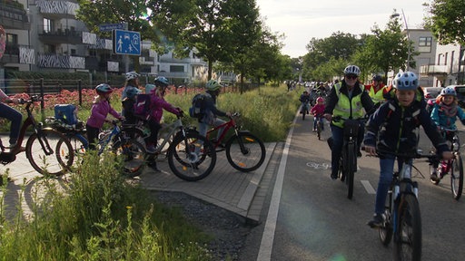 Eine Fahrradkolonne fährt an einer Bushaltestelle vorbei, hier schließen sich weitere Kinder auf ihren Rädern an
