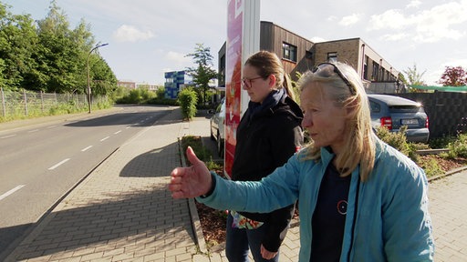 Zwei Frauen stehen auf dem Bordstein und schauen sich die Straße an, die eine Frau zeigt der anderen etwas