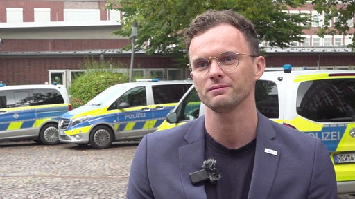 Ein Mann mit runder Brille und dunklem Sakko steht im Vordergrund einer Außenaufnahme. Im Hintergrund sind mehrere blau-gelb markierte Polizeifahrzeuge mit der Aufschrift „POLIZEI“ auf einem gepflasterten Platz vor einem Backsteingebäude zu sehen.