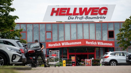 Zu sehen ist der Eingang einer Hellweg-Filiale, ein Mann räumt Einkäufe in sein Auto.