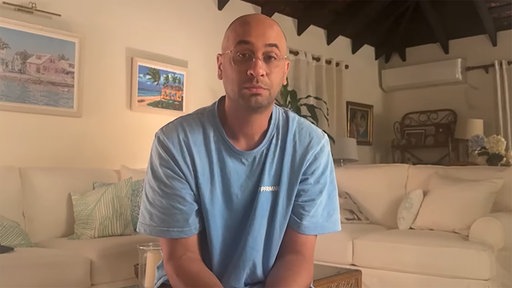 Mann mit Glatze, Brille und blazuem T-Shirt sitzt in einem Wohnzimmer.
