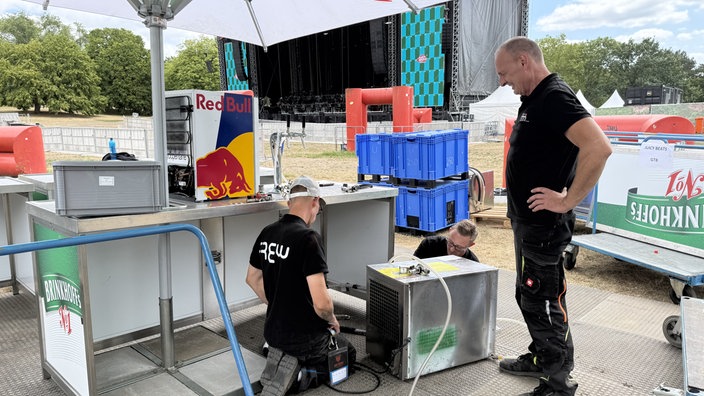 Männer in Arbeitskleidung bauen Getränkeanlage auf Festivalgelände auf.