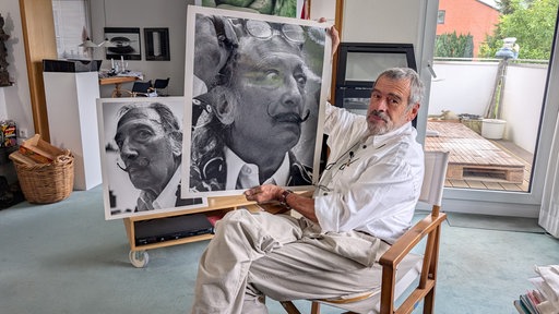 Fotograf Axel Mosler sitzt in einem Raum auf einem Stuhl und hält ein Foto von Dali in die Kamera.