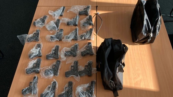Viele in Plastiktüten eingepackte Handfeuerwaffen liegen auf einem Holztisch. Daneben stehen zwei schwarze Taschen.