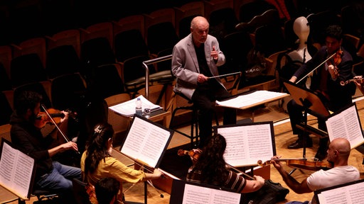 Ein Dirigent gibt in einem Konzertsaal Anweisungen an Musiker, die mit verschiedenen Instrumenten vor Notenblättern sitzen.