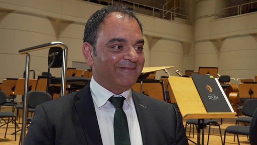 Ein Mann mit dunklen Haaren im Anzug steht in einem Konzertsaal.