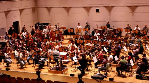 Zahlreiche Musiker, die in einem Halbkreis um ein Dirigentenpult herum sitzen und verschiedene Instrumente spielen.