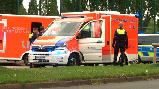 Mehrere Krankentransport Wagen und ein Polizeiwagen stehen auf der Straße, um sie herum stehen Polizisten. 