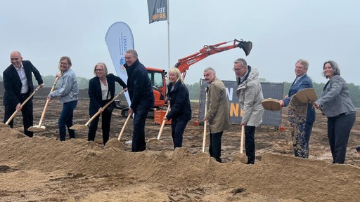 Innenminister Herbert Reul, Polizeipräsidentin Friederike Zurhausen, die Investoren und weitere Beteiligte stehen auf einer sandigen Baustelle und schaufeln Erde für ein Foto