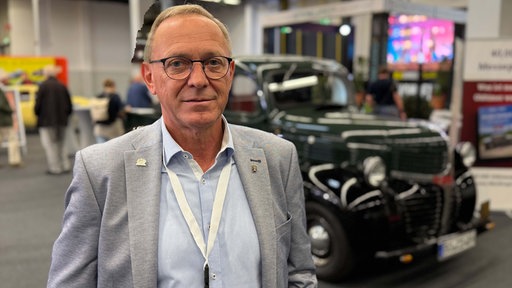 Ein Mann in einem grauen Sakko, der vor einem Oldtimer steht.