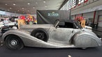 Ein älterer Sportwagen der Marke Mercedes ohne Lack steht in einer Messehalle, das Ersatzrad wird in der Autokarosserie transportiert.