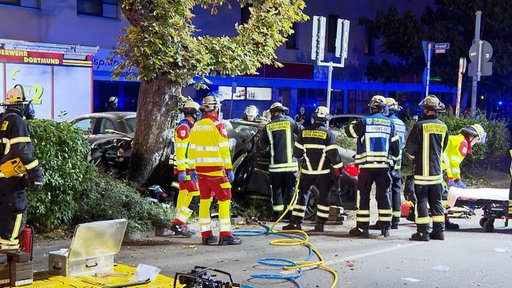 Zehn Einsatzkräfte der Feuerwehr und Sanitäter stehen um ein kaputtes Auto