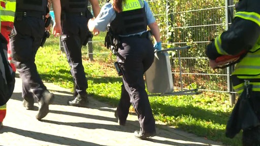 Mehrere Polizisten gehen in eine Richtung, auf dem Bild sind ihre BEine von hinten zu sehen. Sie tragen Uniform. Eine BEamte hält dabei einen Klappstuhl in der Hand.