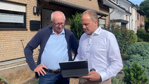Zwei Männer stehen vor einem Einfamlienhaus und schauen in ein Notebook