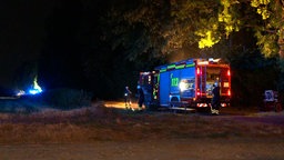 Ein Feuerwehrauto ist von der Seite zu sehen. Es ist Nacht. Zwei Einsatzkräfte stehen neben dem Feuerwehrauto, eine dahinter. Der Brand wird vom Auto verdeckt, Funken steigen auf. 
