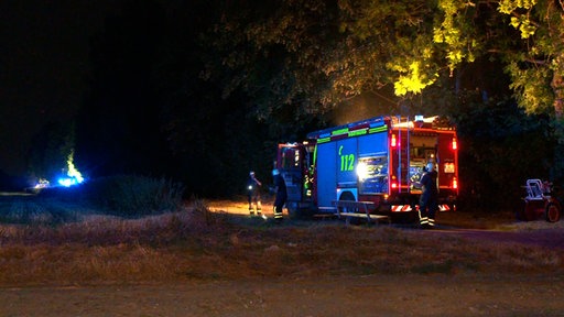 Ein Feuerwehrauto ist von der Seite zu sehen. Es ist Nacht. Zwei Einsatzkräfte stehen neben dem Feuerwehrauto, eine dahinter. Der Brand wird vom Auto verdeckt, Funken steigen auf. 