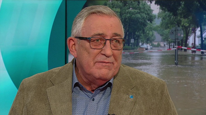 Ein älterer Mann mit Brille, in Hemd und Jackett vor einem Monitor-Hochwasserbild
