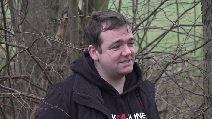 Ein junger Mann mit schwarzem Hoodie, schwarzer Jacke vor mehreren Bäumen