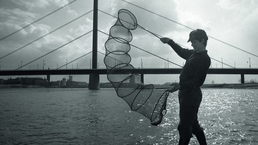 Ein Mann hält ein langes Netz, einen Setzkescher. Unten im Netz ist ein Fisch. Er steht am Rhein, im Hintergrund ist eine Brücke sichtbar.