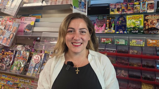 Eine Frau steht vor einem Kioskregal und lächelt in die Kamera