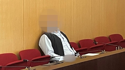 Der Angeklagte sitzt auf der dunkelroten Anklagebank. Er trägt ein hellblaues Hemd und eine schwarze Weste. Der Kopf ist zum Identitätsschutz verpixelt.