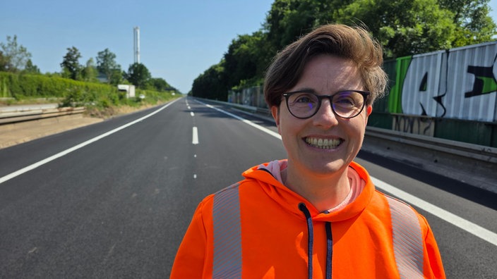 Eine lachende Frau mit runder Brille und kurzen Haaren und neonorgangenem Pulli auf einer leeren Autobahn