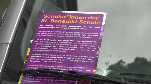 Ein lilaner Flyer mit gelber Schrift ist unter dem Scheibenwischer eines Autos geklemmt