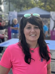 Eine Frau mit einem pinken Tshirt lächelt in die Kamera. Im Hintegrund spielen zwei Frauen Tischtennis.