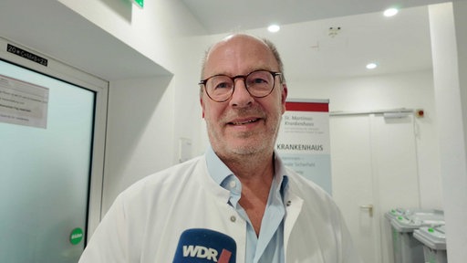 Ein Mann mittleren Alters trägt einen weißen Kitten und eine brille und lächelt in die Kamera. Er hat lichtes Haar.