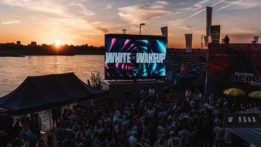 Viele Menschen vor Leinwänden am Rheinufer, im Hintergrund der Sonnenuntergang