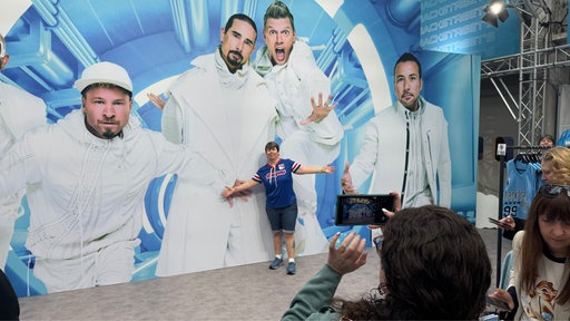 Person posiert mit ausgebreitreten Armen vor Fotowand. Fotowand zeigt Member der Backstreet- Boys.