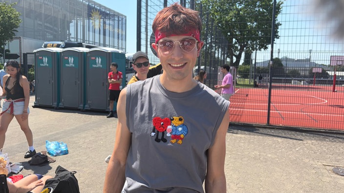Ein junger Mann mit Sonnenbrille steht in der Sonne vor der Merkur-Spiel-Arena. Er trägt ein besticktes T-Shirt.