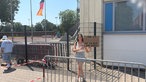 Eine junge Frau steht in einem abgesperrten Wartebereich und hält ein Plakat hoch, auf dem ein spanischer Spruch steht.
