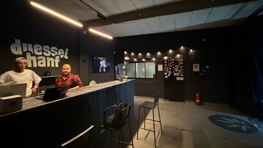 Zwei Männer stehen hinter einer Theke im Raum eines Cannabis Social Clubs