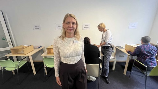 Eine blonde Frau mittleren Alters lächelt leicht in die Kamera. Im Hintegrund sitzen zwei Frauen.