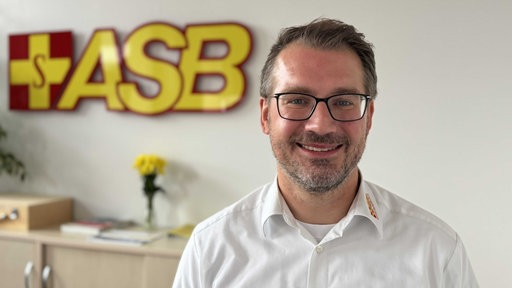 Ein Mann mit Brille lächelt in die Kamera, hinter ihm sind gelbe Buchstaben zu sehen, sie bilden das Logo "ASB"