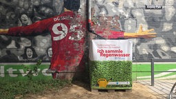 Der Regenwasser-Container am Kolpingplatz in Düsseldorf. Er steht vor einer bemalten Wand.