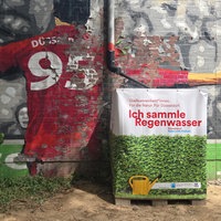 Der Regenwasser-Container am Kolpingplatz in Düsseldorf. Er steht vor einer bemalten Wand.