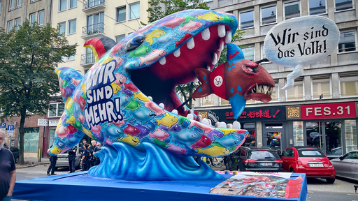 Ein Mottowagen auf der Demo, dargestellt ist ein großer Fisch der einen kleineren frisst, der größere beschriftet mit "Wir sind mehr", der keiner mit "Wir sind das Volk"