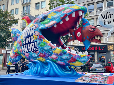 Ein Mottowagen auf der Demo, dargestellt ist ein großer Fisch der einen kleineren frisst, der größere beschriftet mit "Wir sind mehr", der keiner mit "Wir sind das Volk"
