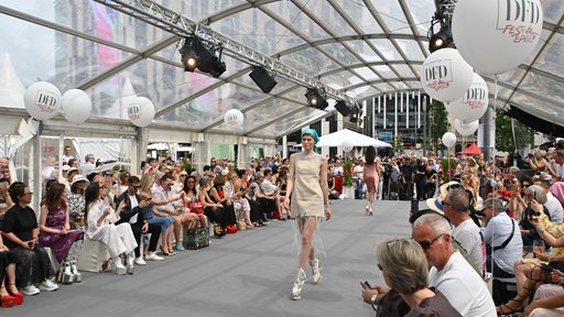 Eine Frau in einem Kleid geht über einen Laufsteg. Rechts und links sitzen viele Zuschauer.