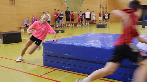 Sporthalle in einer Schule. Kinder laufen in Schräglage um Matte.
