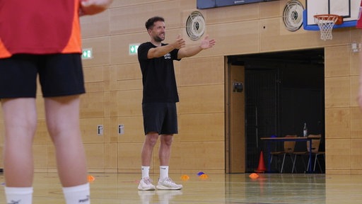 Sporthalle in einer Schule. Sportlehrer zeigt mit Armen Übung an.