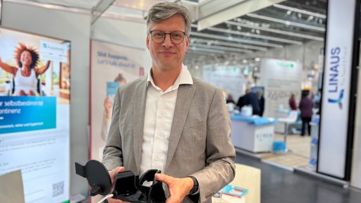 Professor Urs Schneider steht mit Hemd und grauem Sakko an einem Stand auf der Rehacare in Düsseldorf